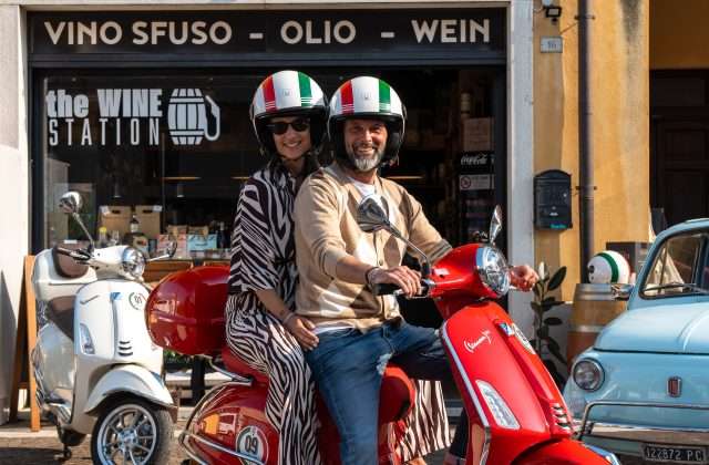 Coppia in Vespa rossa con caschi tricolore davanti a The Wine Station di RentGarda a Bardolino, shop di vino sfuso e olio